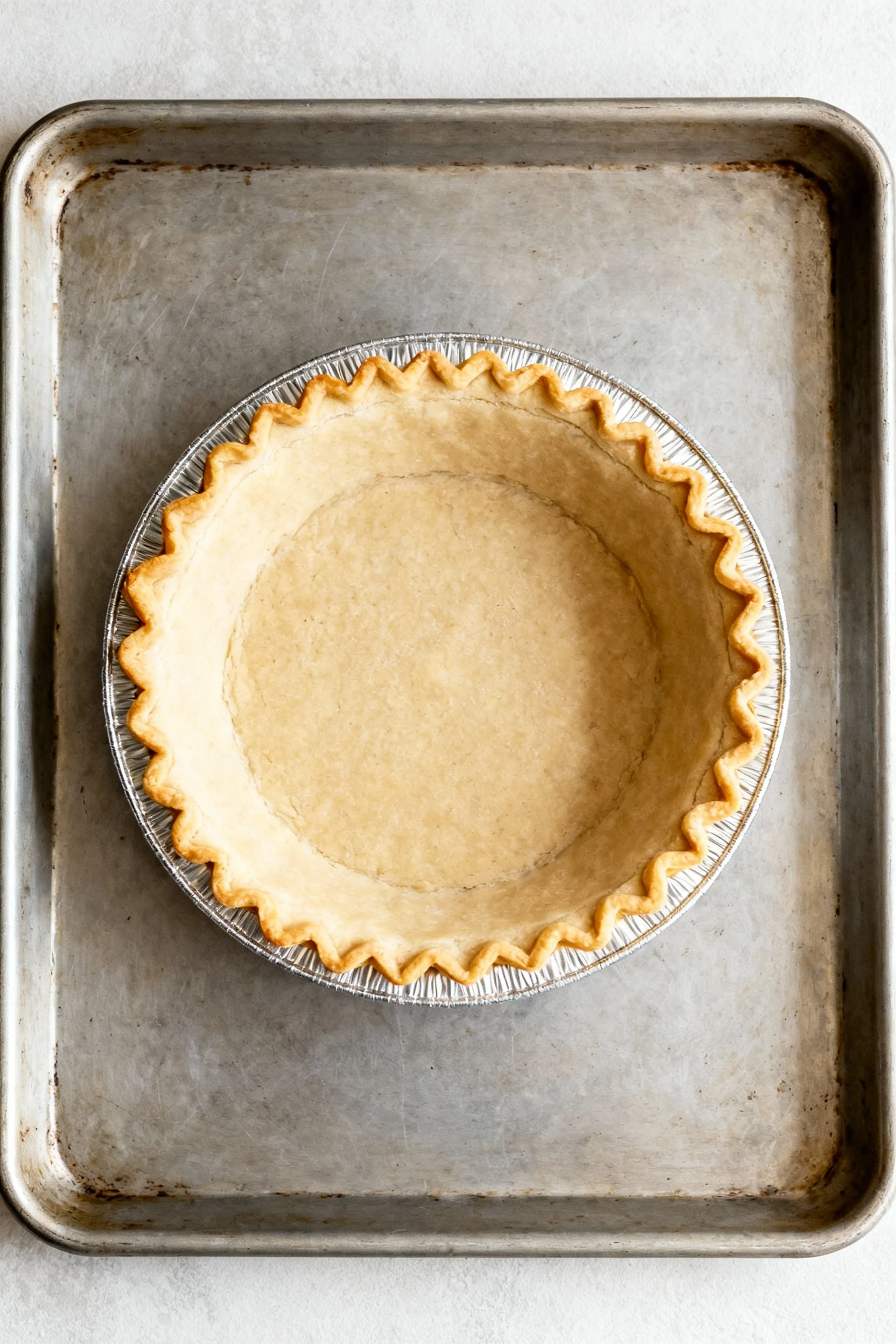 2. Overhead of the parbaked 9-inch deep-dish pie crust in its foil tin on a preheated rimmed sheet pan, crimped rim gold