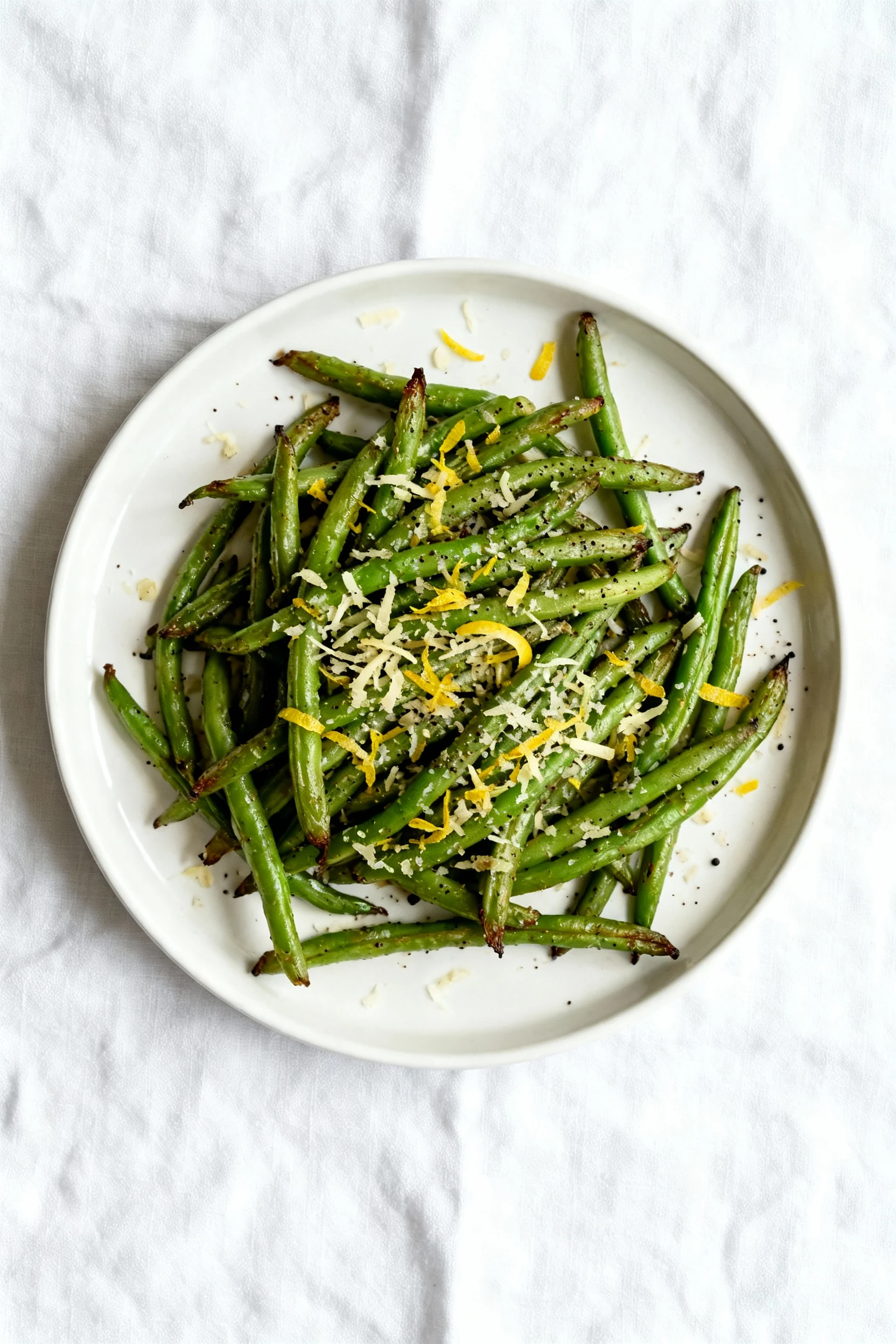 Overhead shot of garlic–Parmesan air fryer green beans piled on a matte white plate, golden cheese flecks, lemon zest fi