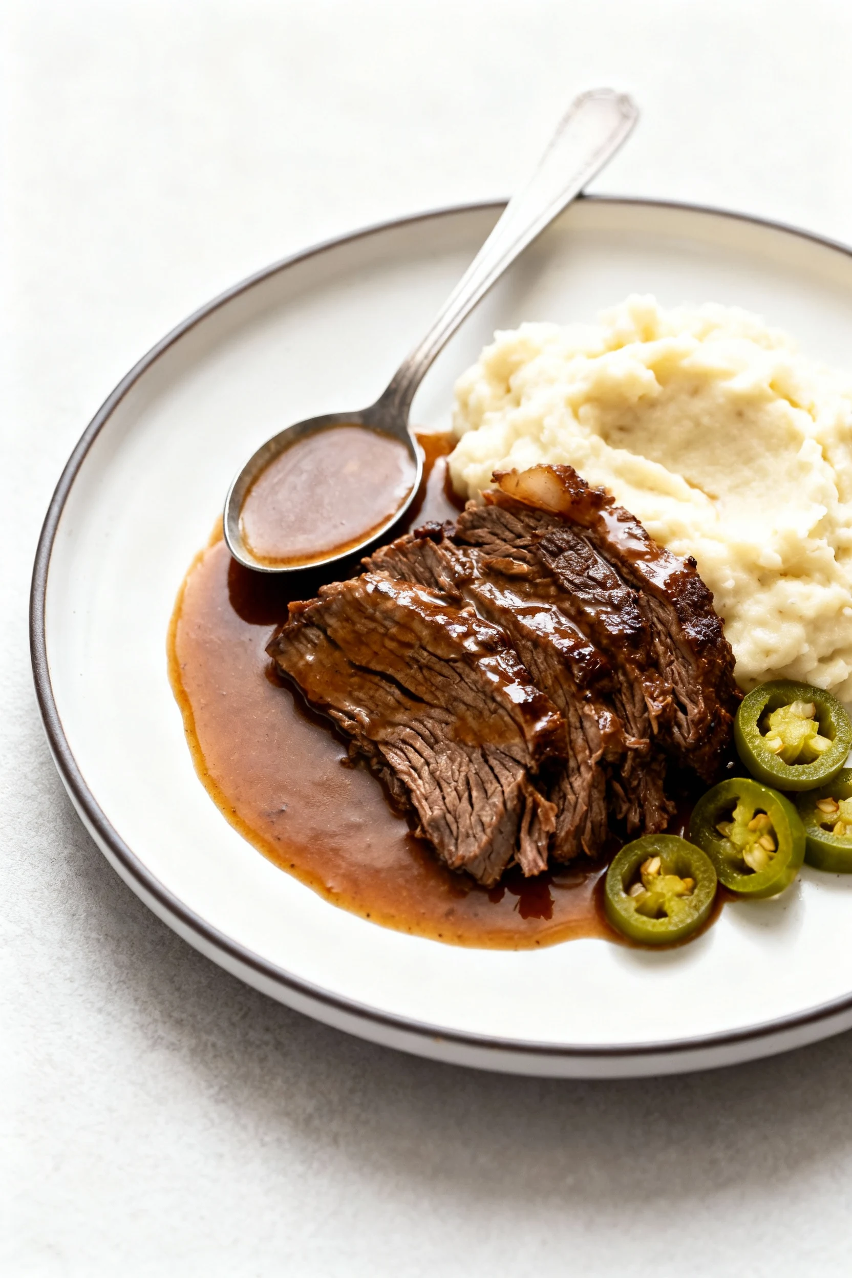 Beautifully plated Mississippi pot roast over creamy mashed potatoes on a white rimmed plate: generous spooned jus pooli