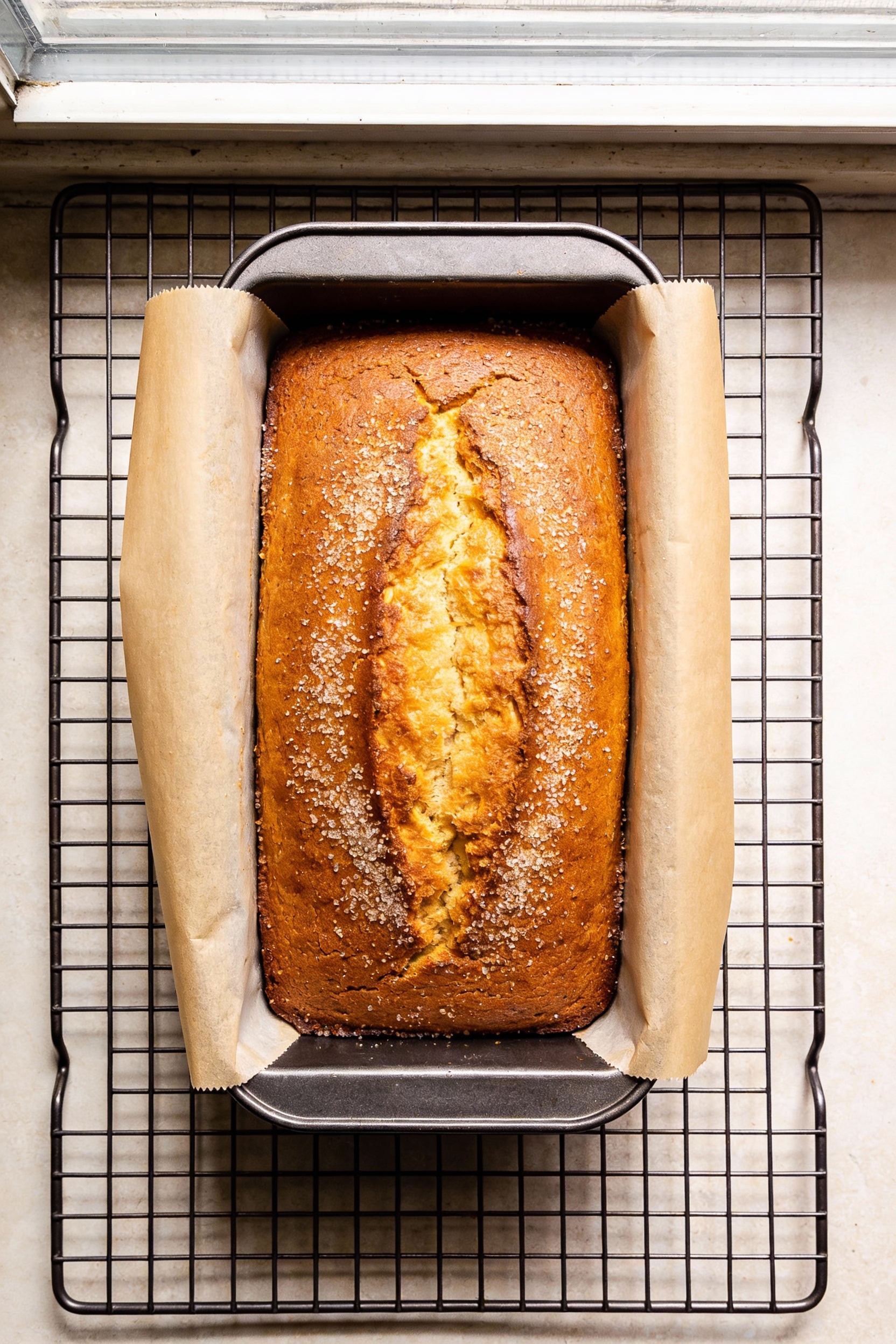 Overhead shot of a freshly baked loaf cake on a wire rack, lifted with a parchment sling from a 9x5-inch pan; golden-bro