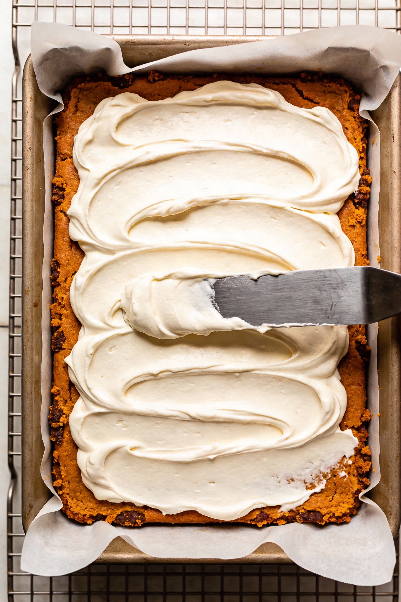 Cooking process: Pumpkin sheet cake just cooled on a wire rack, offset spatula creating silky cream cheese frosting swir