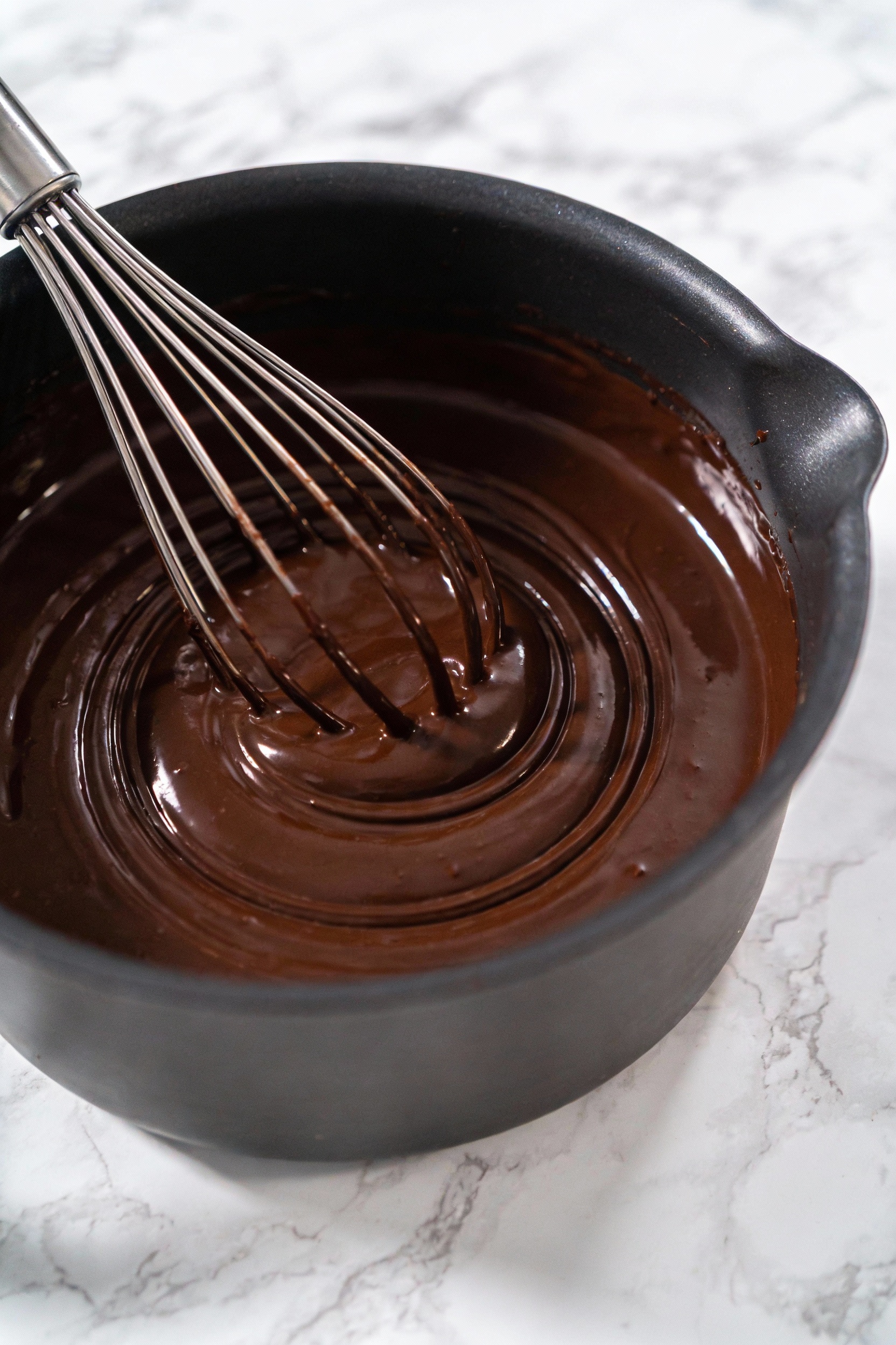 Dark chocolate ganache mid-whisk in a heatproof bowl—concentric ripples forming from the center, ultra-glossy 60–70% cho
