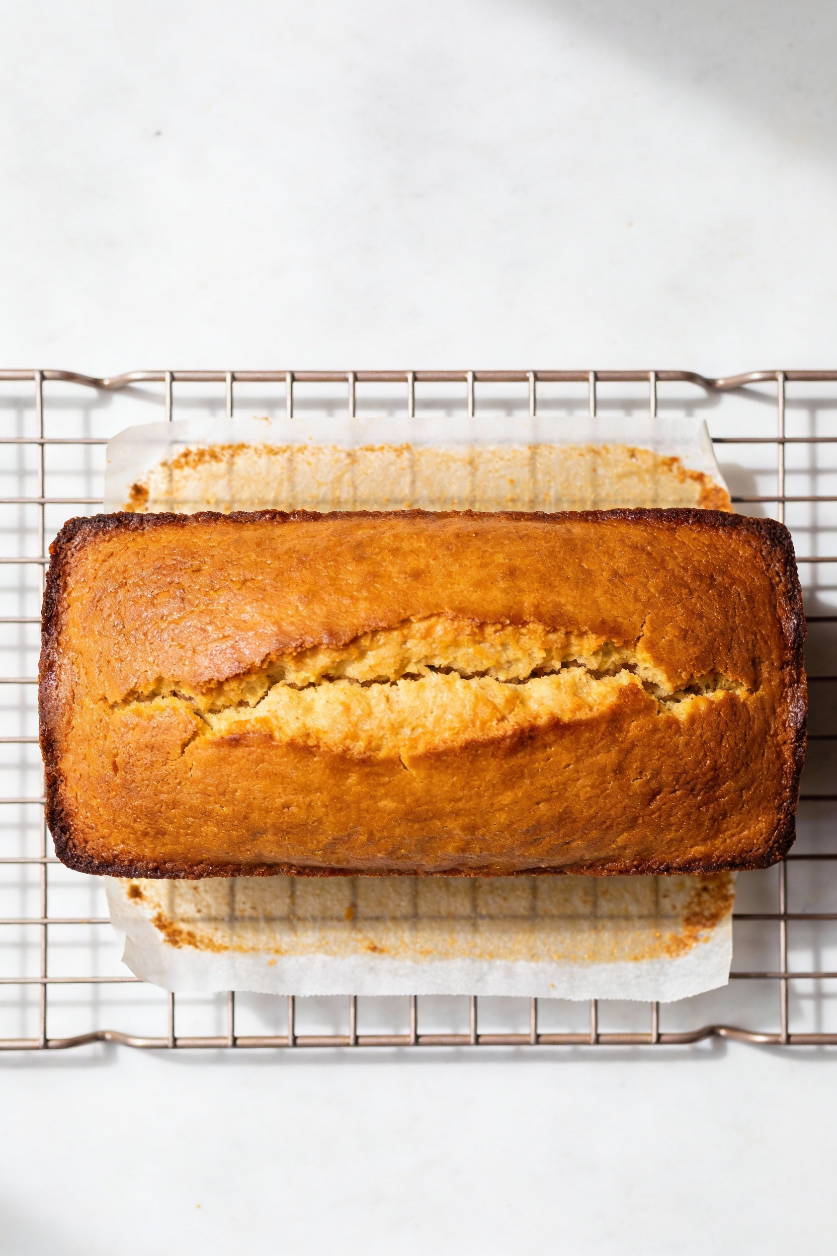 Overhead shot of the whole deep-golden pound cake loaf on a cooling rack with parchment sling visible, signature crack d