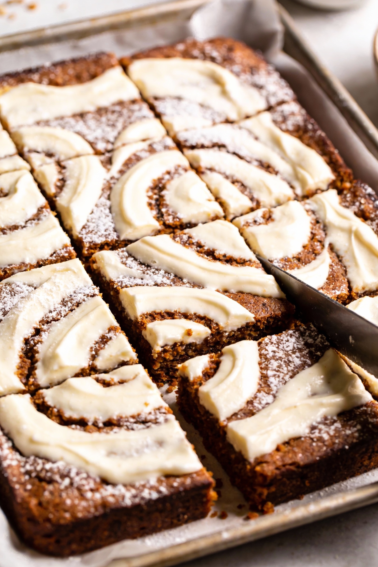 Overhead shot of cream cheese swirl bars on parchment-lined metal 9x13 pan, sharp knife-cut swirls of creamy white again