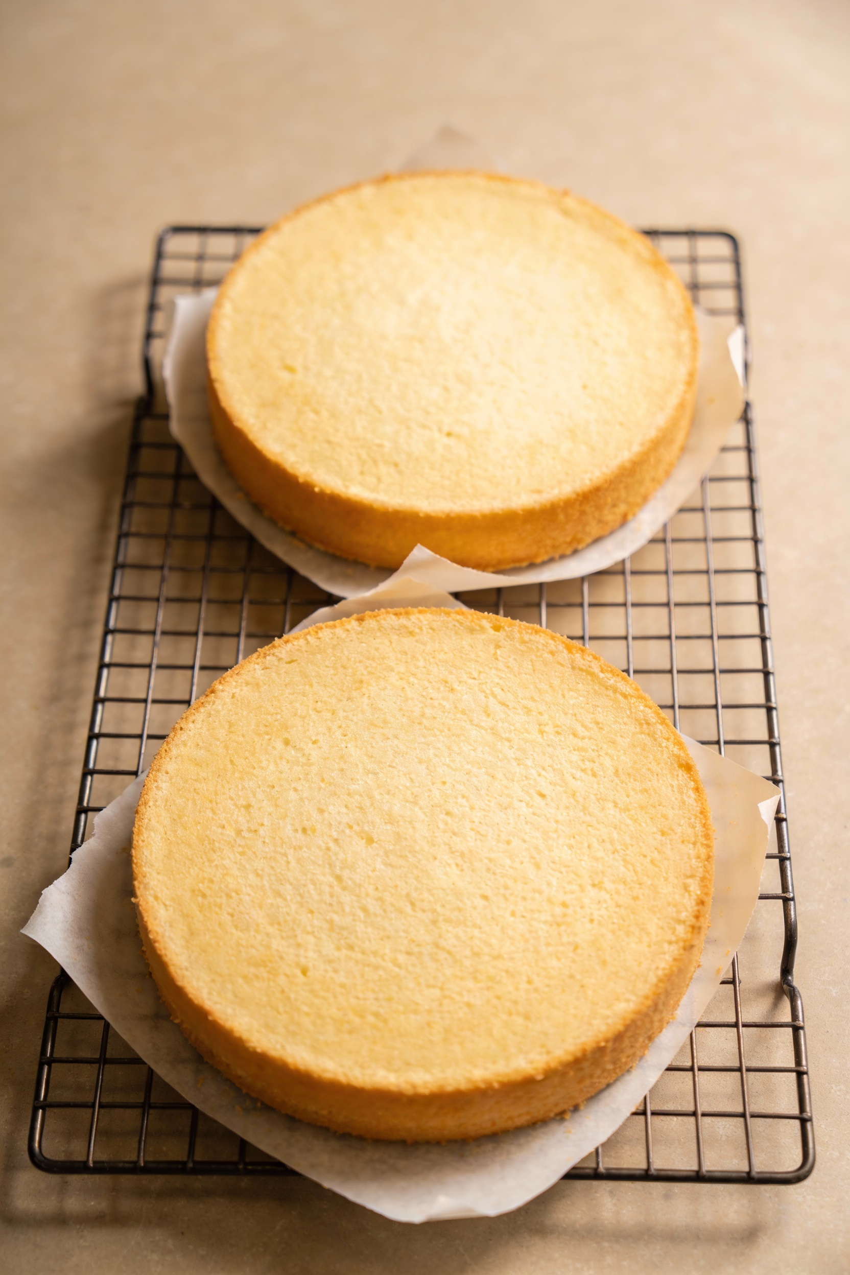 Cooking process: two baked 8-inch vanilla cake layers cooling on a wire rack, parchment paper peeled back at the edges, 