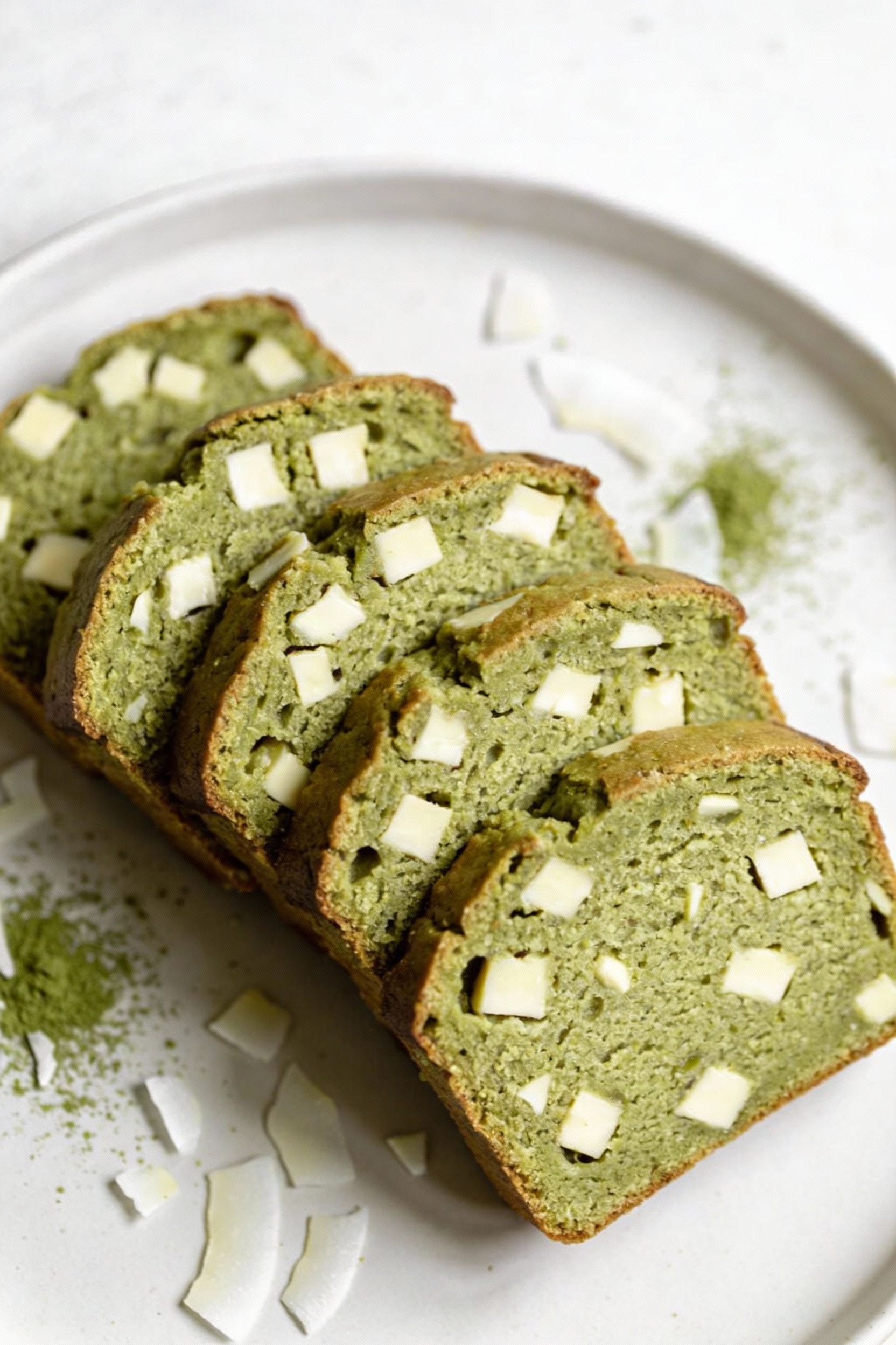 Beautifully plated matcha white chocolate loaf: several neat slices fanned on a matte white plate; pale-green crumb stud