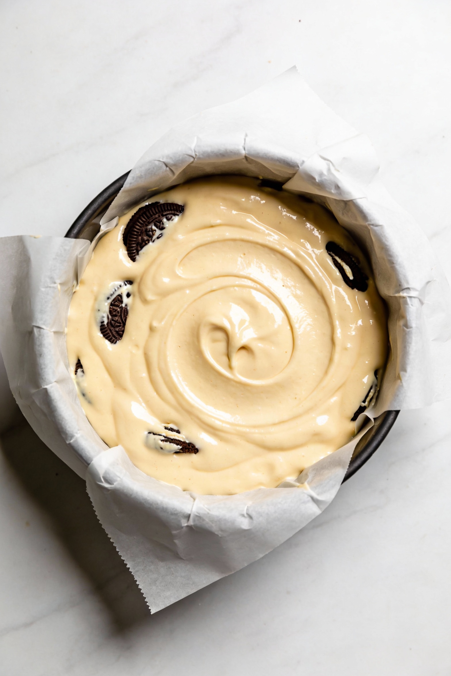 Cake batter resting in a parchment-lined round pan before baking, thick and glossy pale batter with visible cookie piece