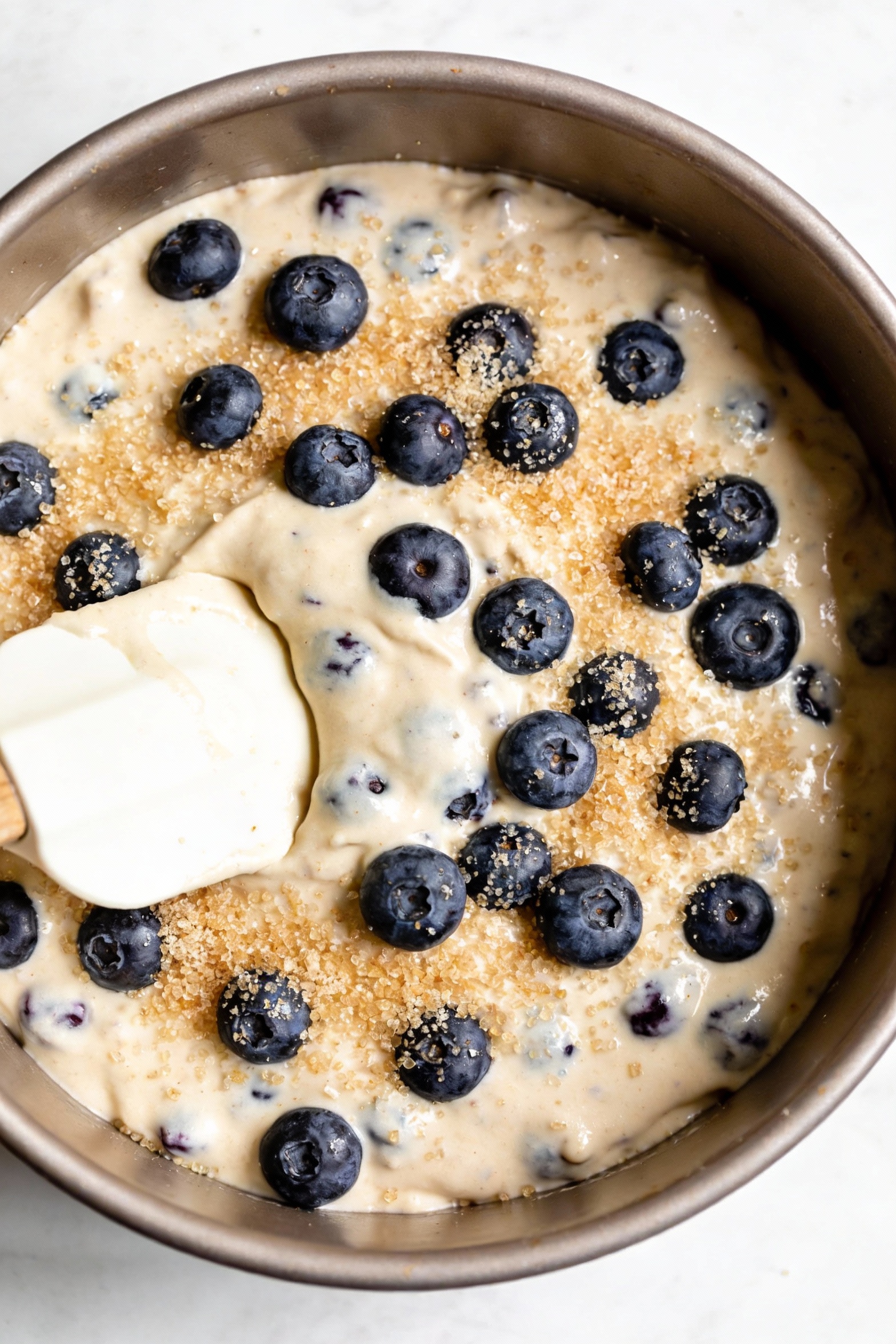 Cooking process: blueberry cake batter in a round pan mid-prep, thick batter smoothed with a spatula, extra blueberries 