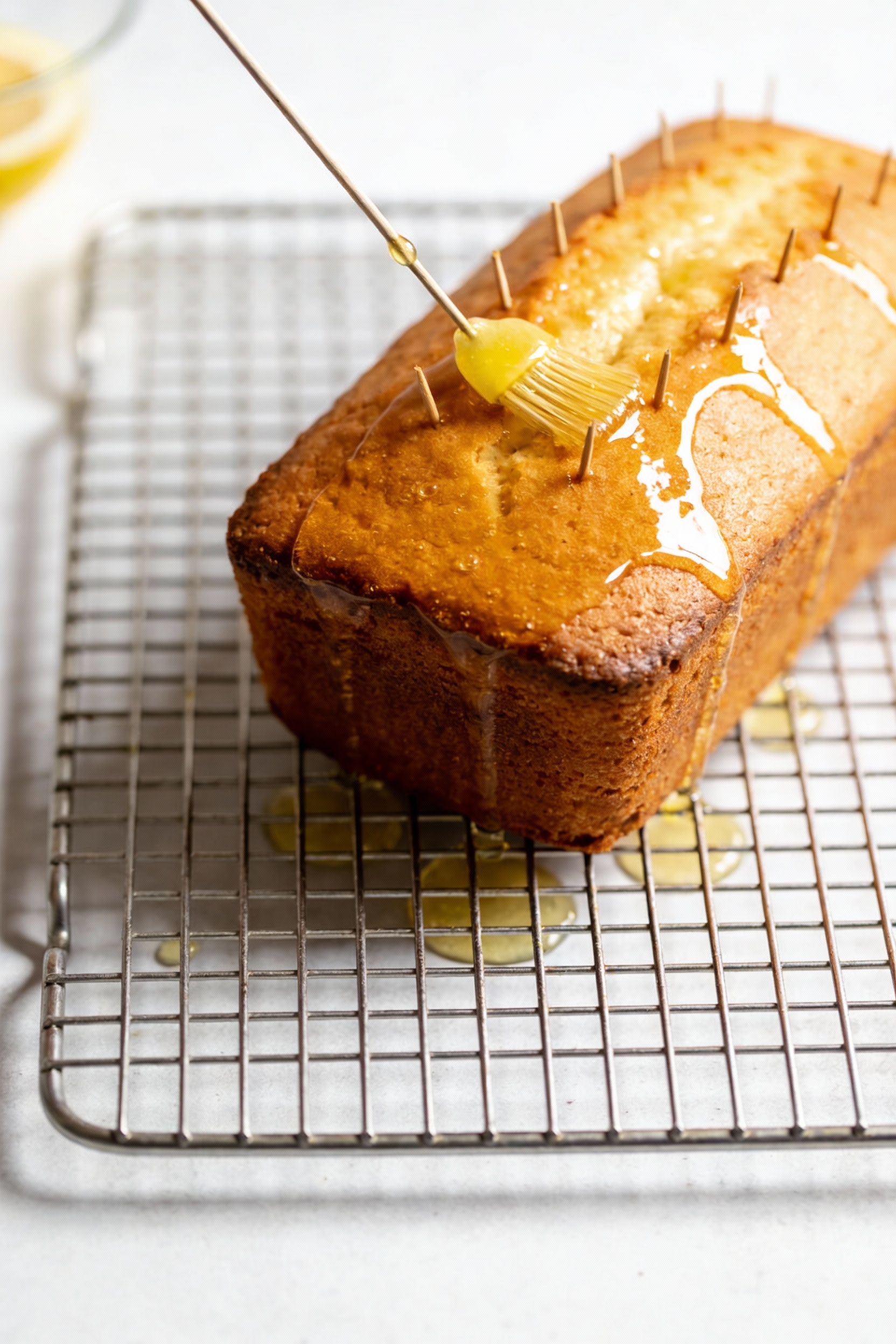 4. Cooking process: warm lemon cake on a cooling rack being poked evenly with a skewer, then brushed with a shiny lemon 