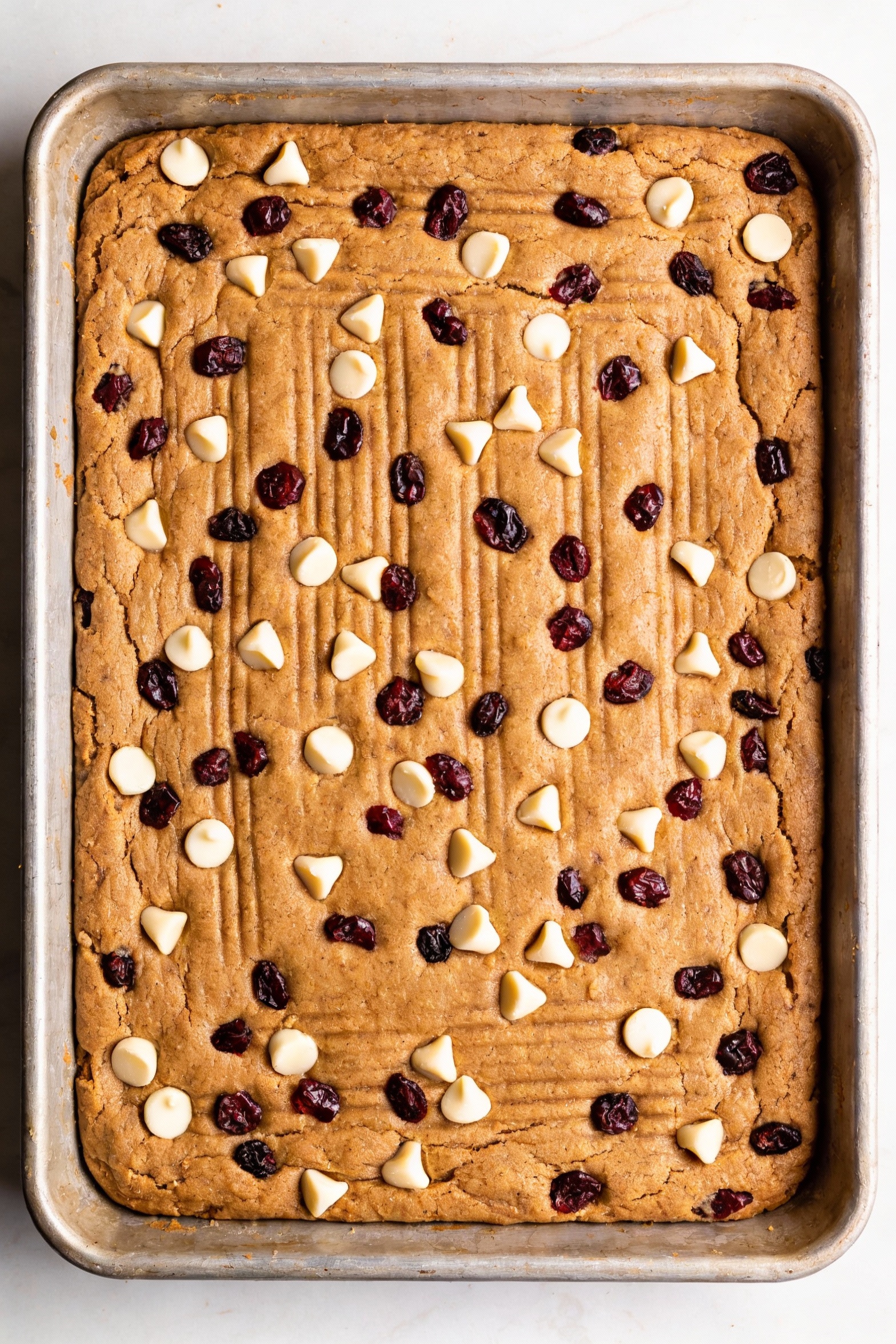 Cooking process: thick spiced cookie bar dough being pressed evenly into a greased 9x13 pan, dough studded with white ch